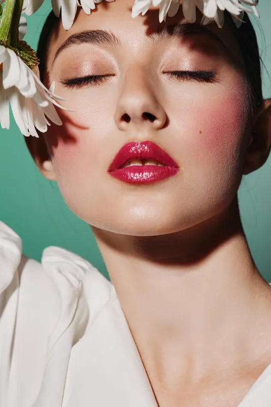 Close-up of a woman with glossy red lips and soft pink makeup wears a white top with closed eyes, surrounded by white flowers. Concept of beauty, creative, fashion.