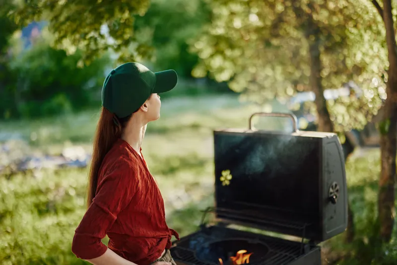 Young woman in a green cap enjoying a summer barbecue outdoors, surrounded by trees and greenery, showcasing a joyful attitude and a sense of relaxation
