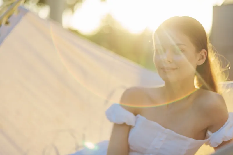 Beautiful young woman in a white off shoulder dress, enjoying sunshine in a serene outdoor setting, conveying warmth, happiness, and tranquility during summer