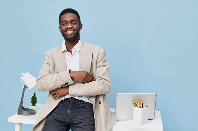 Portrait of a smiling young African American man in casual blazer sitting at modern desk with laptop and stationery, looking confidently at camera. Studio shot with bright pastel background. People li