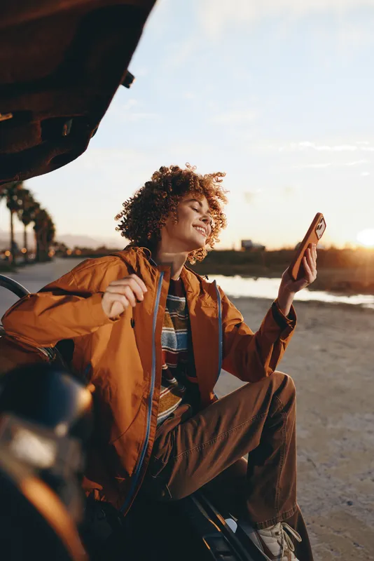 Woman lifestyle at beach sunset, smiling with smartphone for selfie, curly hair and jacket near car, enjoying roadtrip moment, casual travel outdoors in warm glow.
