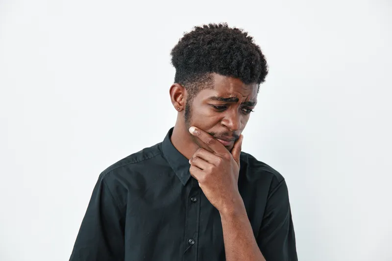 Young black man with dark skin and curly hair wearing black shirt looking down with thoughtful expression on white background. Portrait of pensive african male thinking.