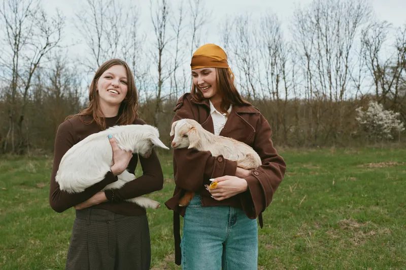 Two women outdoors in a rural field hold young goats, smiling warmly, casual jackets, gentle interaction, natural sunlight, lifestyle moment, rustic ambiance, soft connection with animals