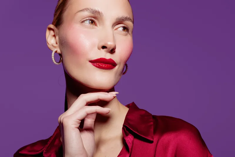 Portrait of confident woman with red lipstick and shiny skin wearing red satin blouse and hoop earrings on purple background. Concept of beauty, fashion, and elegance.