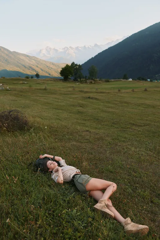 Calm portrait of a woman resting in a meadow beneath distant hills, enjoying outdoor space, sunlit grass and tranquil scenery, a quiet moment of leisure and nature.
