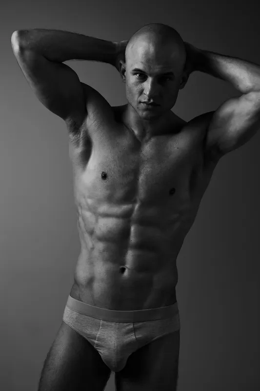 A strong man posing confidently in black and white, showcasing his toned physique and expressing strength and determination in a minimalist setting.