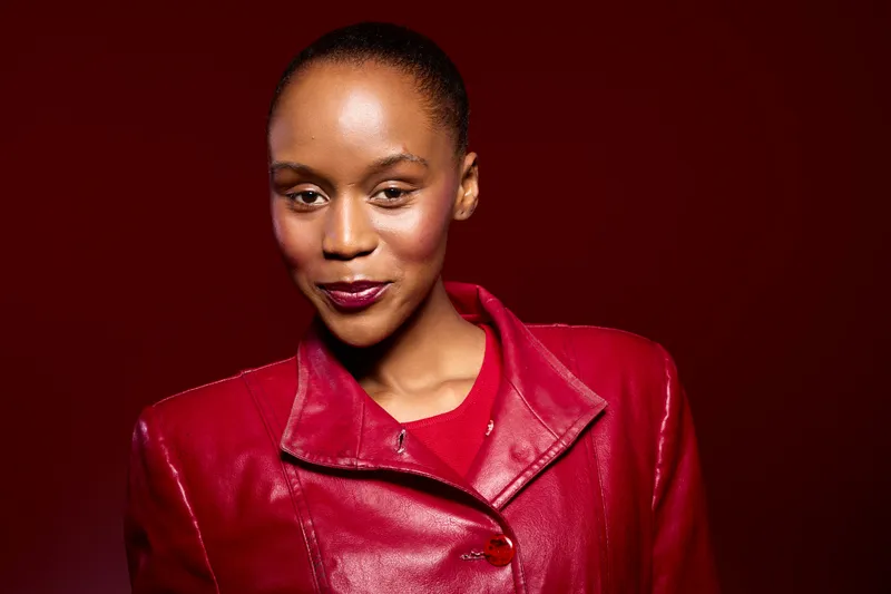 Confident African woman with dark skin wearing red leather jacket smiles softly against deep red background. Portrait of beauty, fashion, and elegance with vibrant color contrast.