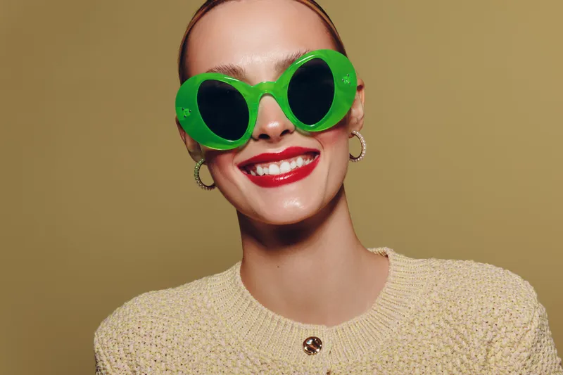 Smiling woman with bright red lipstick wearing large green sunglasses and gold hoop earrings. She wears a beige knitted sweater against a plain brown background. Fashion and style concept.
