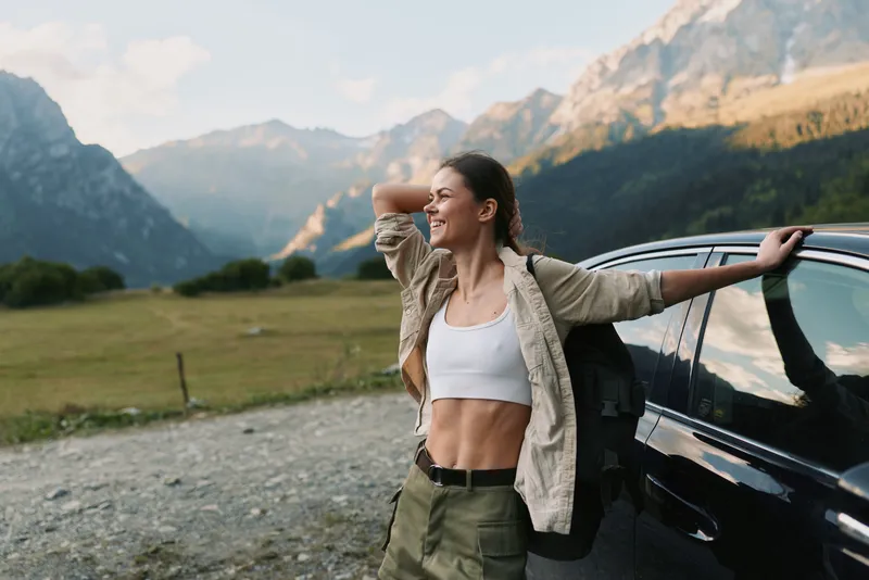 Smiling woman outdoors leaning on car door, casual outfit, relaxed posture, mountain landscape background, sunny day, natural light, lifestyle concept