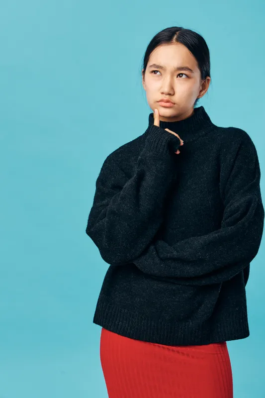 Young woman in black sweater and red skirt stands thinking with hand on chin against blue background, portrait of pensive female in casual outfit with thoughtful expression.