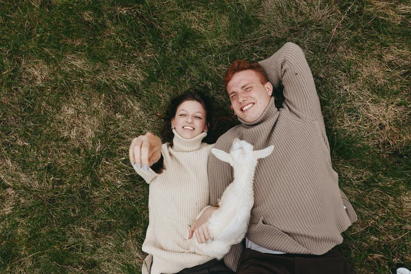 A couple lying on grass outdoors with a curious goat kid, smiling and relaxed, showcasing cozy lifestyle, playful moment, and gentle bond under warm sunlight.