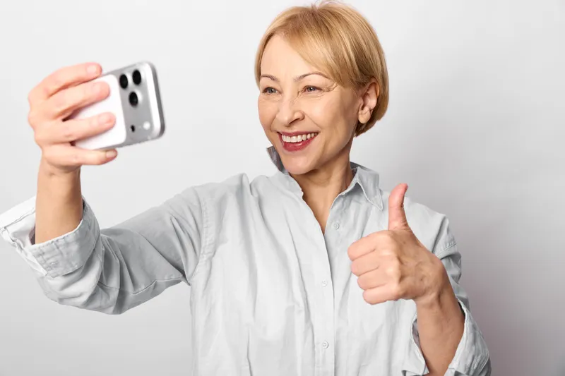 Woman selfie with smartphone, with smile, giving thumbs up, older adult indoors, soft light, neutral background, natural skin tones, casual shirt, bright