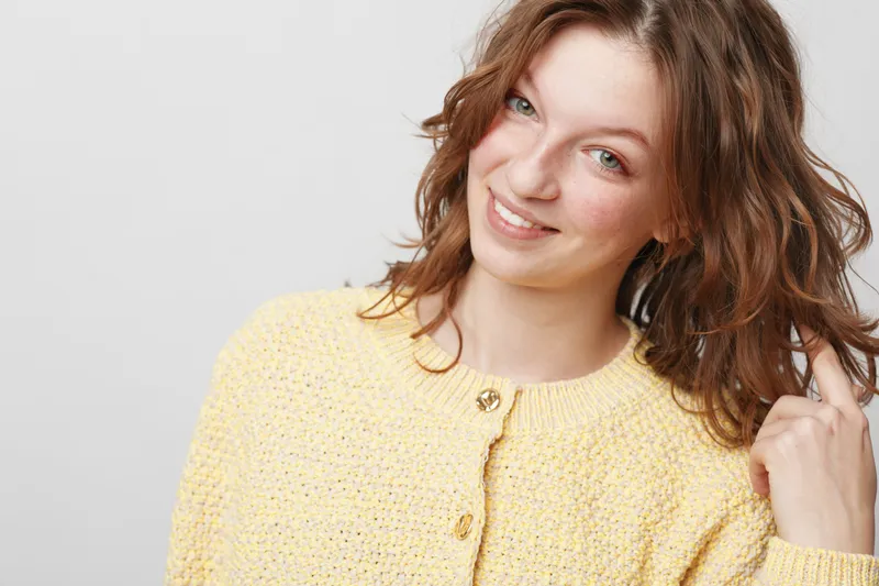Smiling young woman with wavy brown hair wearing a yellow knitted sweater looks at camera with a friendly expression. Studio portrait on light grey background. Concept of happiness and casual style.