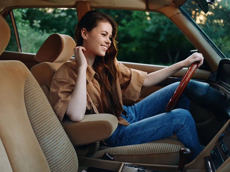 Young woman smiling in vintage car interior, wearing casual brown shirt and blue jeans, with lush green background, embodying freedom and joy.