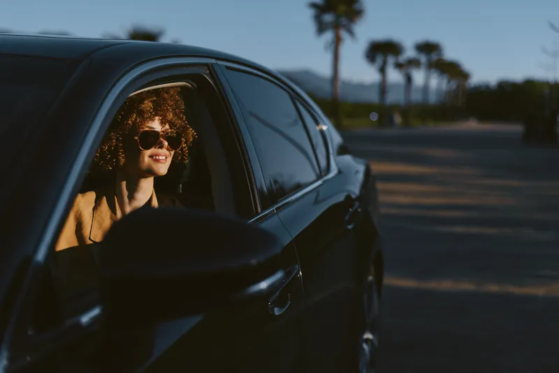 Candid woman in a sleek car, sunlit smile, outdoors during a relaxed drive, retro film color palette, warm tones, casual style, palm trees in the background, natural lighting