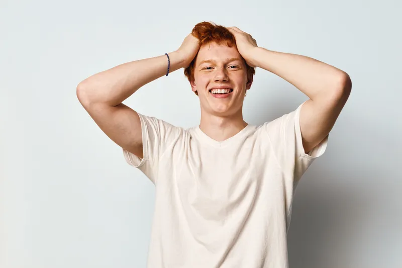 Young man with red hair smiling and holding head with hands on white background. Casual style, happy expression and relaxed male in comfortable t shirt portrait.