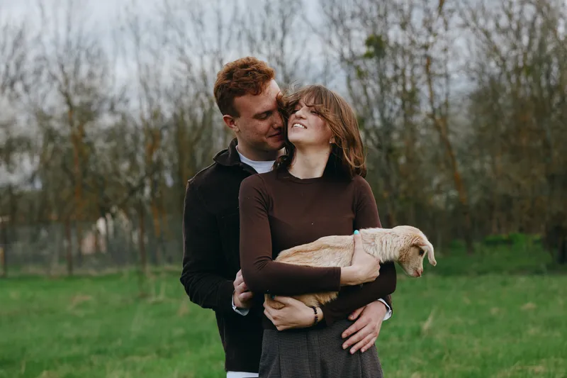 Couple outdoors in a park embrace the smiling woman who holds a goose, while the man wraps his arms around them, a warm lifestyle moment of affection and gentle animal companionship today