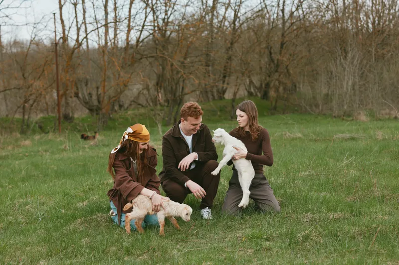 A family gathers in a grassy field with a small kid goat and a curious dog, sharing a gentle moment outdoors. A lifestyle portrait capturing bond, playfulness, and rural life, with children and