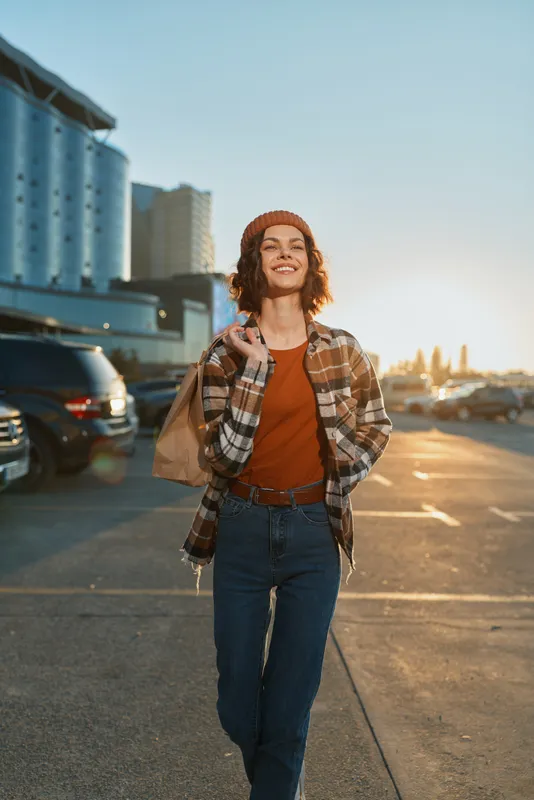 Woman smile walking in urban parking lot at golden hour carrying a paper bag, candid lifestyle portrait emphasizing authenticity, mindful living and emotional storytelling with warm glow.
