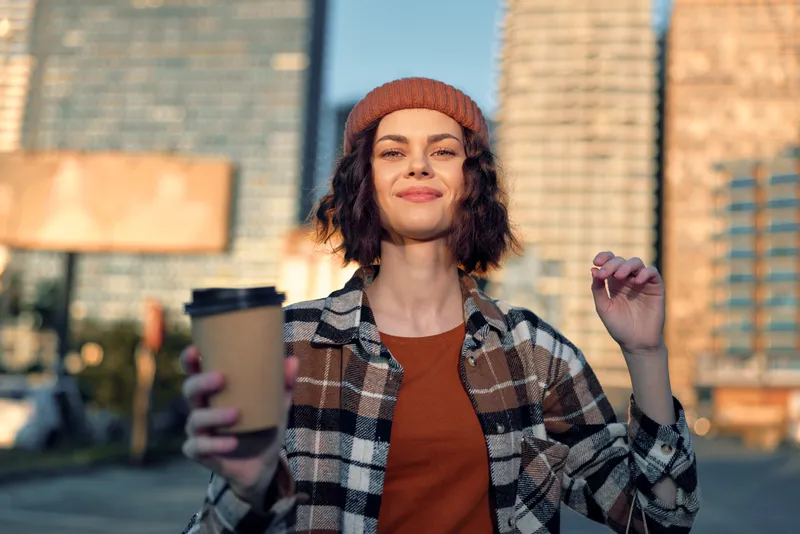 woman coffee urban smile beanie sunlight: Young woman holds a takeaway coffee on a city street at golden hour glow, candid lifestyle portrait conveying authenticity, mindful living and emotional