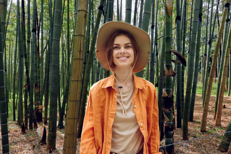 Woman in a bamboo forest wearing a wide hat and orange shirt, smiling in portrait style among green trees and nature, outdoors scene conveying joy and peaceful exploration.