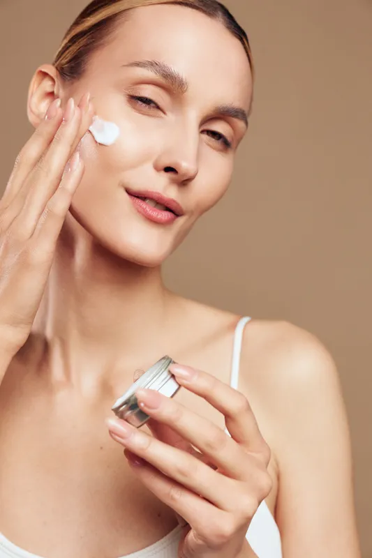 Young woman applies moisturizing cream on her face with fingers holds small jar skincare beauty routine natural skin care closeup isolated on beige background