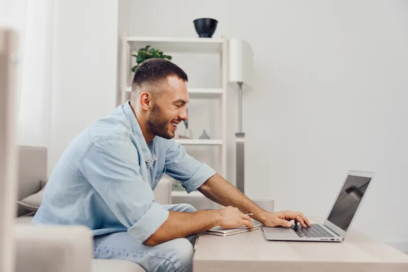 Focused man engaging with a laptop, showcasing modern home office style while wearing casual attire. The setting is bright and minimalistic, creating a productive atmosphere.