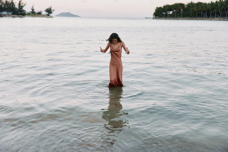 A woman in a soft pink dress stands in shallow turquoise water, gentle waves curling around her ankles, calm horizon, peaceful coastal moment, reflective surface.