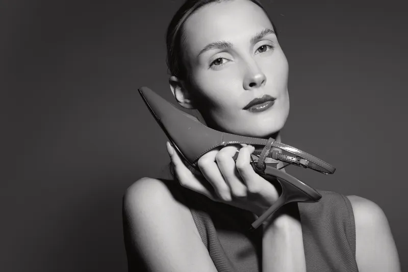 Elegant Caucasian woman with pale skin holds stylish high heel shoe near face, looking confidently. Black and white artistic portrait with fashion concept and minimal studio lighting.