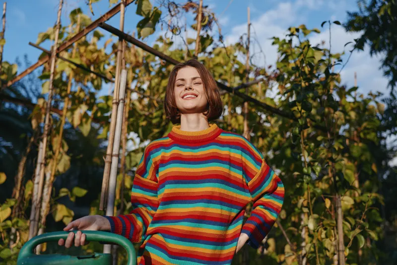 Woman smile in colorful striped sweater in garden holding a watering can among green plants and trellis, outdoor portrait of happy gardener enjoying a sunny day in nature.