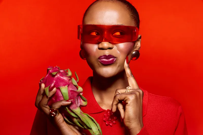 Stylish dark skinned African woman wearing red sunglasses and red sweater holds dragon fruit, posing against vibrant red background. Concept of fashion, exotic fruit, and modern beauty.