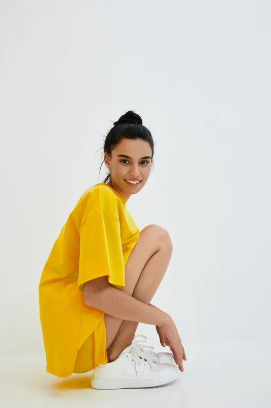 smiling woman wearing oversized yellow t-shirt and white sneakers squatting on white background with casual hairstyle, expressing relaxed and cheerful mood