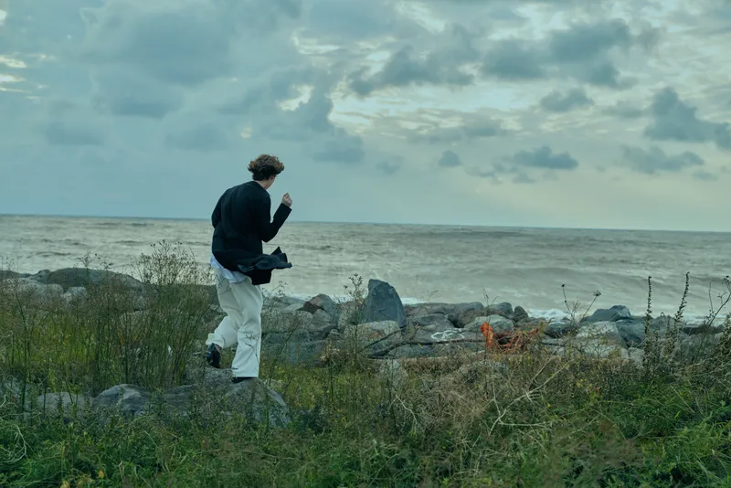 Man walking along a rocky coastline with a camera bag under a cloudy sky, rugged shoreline and calm ocean creating a moody outdoor landscape