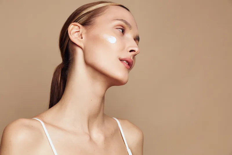 Young woman with clear skin applying white cream on cheek, side profile, natural makeup, smooth complexion, minimal beige background, skincare routine concept