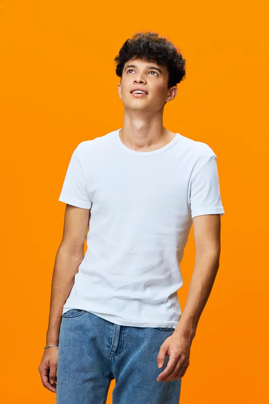 young man wearing a white t-shirt and blue jeans stands confidently against a bright orange background, looking upwards with a thoughtful expression