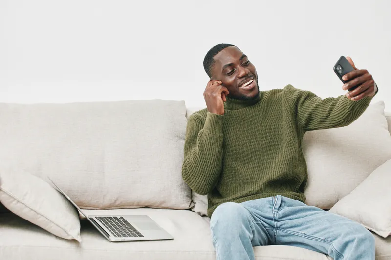 Smiling Black man in a green sweater enjoying video call on smartphone, relaxing on couch with laptop, modern home office setting