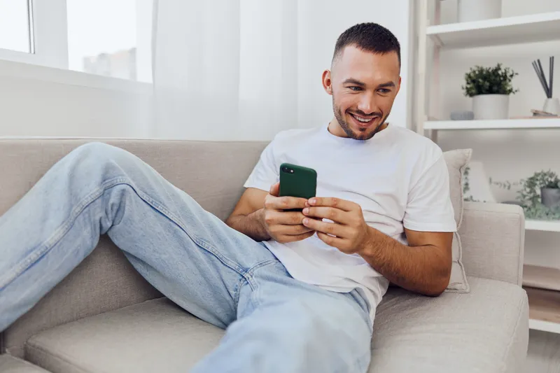 Smiling man relaxing at home, using smartphone with a joyful expression. Bright lighting adds warmth to the cozy atmosphere.