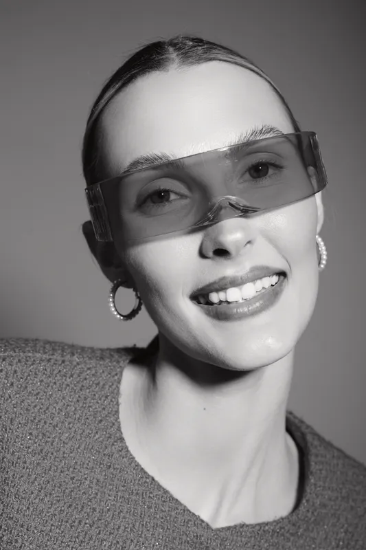 Smiling white woman with fair skin wearing oversized transparent glasses and hoop earrings looks at camera in studio. Concept for fashion and portrait.