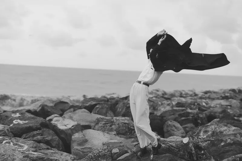 Freedom, movement and wind shape a figure on a rocky coast, cloak flaring, outdoors by the sea. A dynamic, monochrome moment of release and energy, suitable for creative stock scenes.