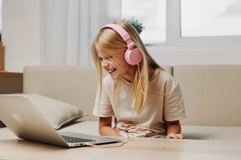 Happy girl with long blonde hair wearing pink headphones laughs while watching laptop in cozy living room, natural light from window brightens warm home interior.