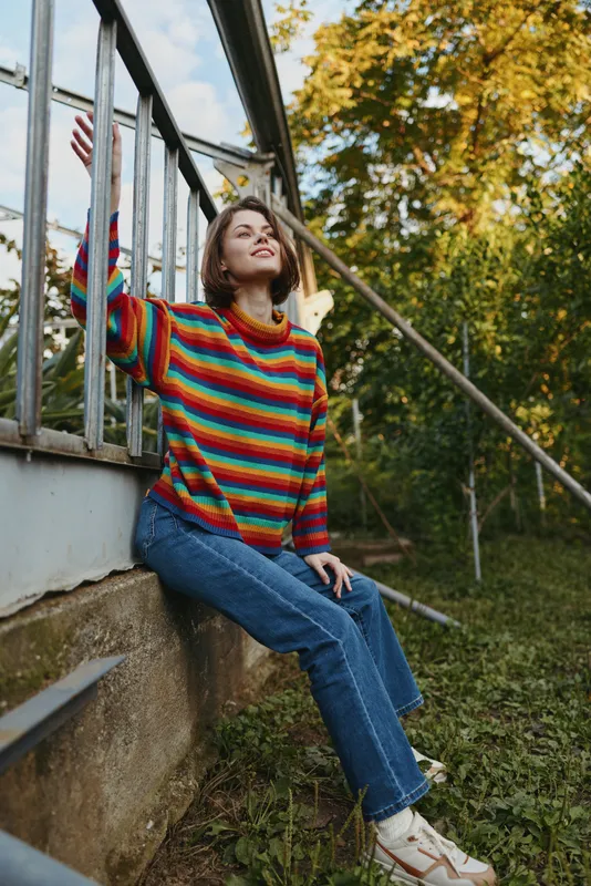 woman sweater jeans outdoors smile railing, young adult sitting on concrete ledge by metal fence, colorful striped knit jumper with denim and sneakers, relaxed autumn park portrait