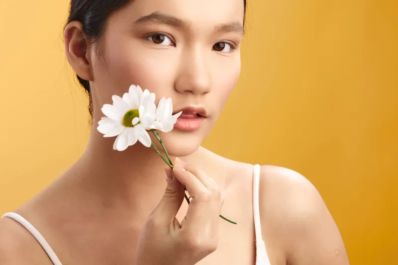 Asian woman holds a white daisy near her lips, looking softly at the camera against a warm yellow backdrop. The scene conveys delicate beauty, calm mood, and graceful charm.