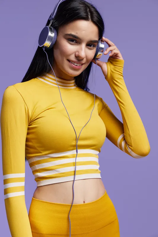 Woman sport fitness wearing yellow athletic outfit and headphones, smiling confidently against a purple background, showcasing healthy lifestyle and active spirit.