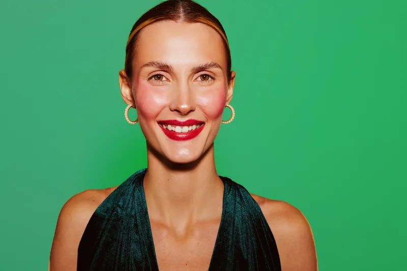 Smiling woman with red lipstick and gold hoop earrings poses against green background. She wears a dark sleeveless top and has natural makeup with rosy cheeks. Portrait concept.