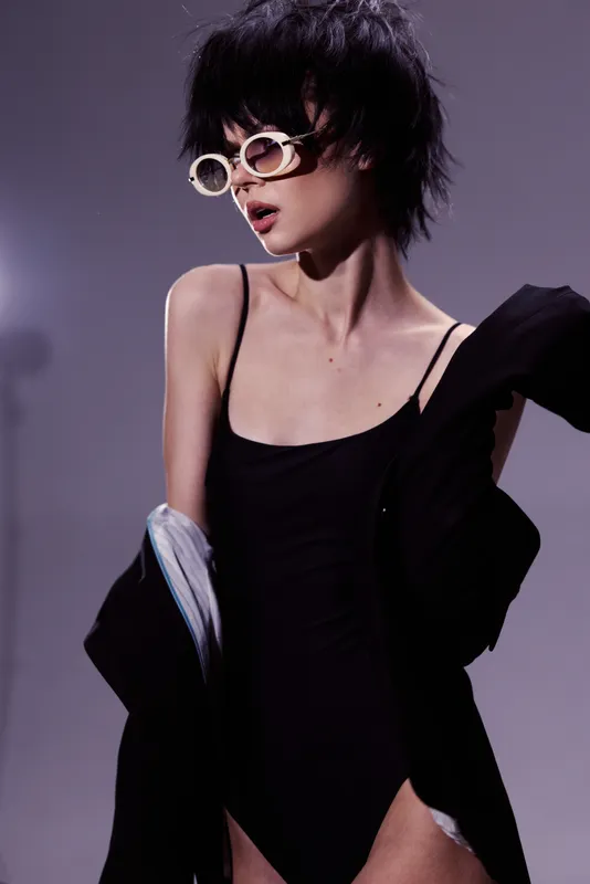 Stylish woman wearing black bodysuit and white sunglasses posing with jacket on purple background