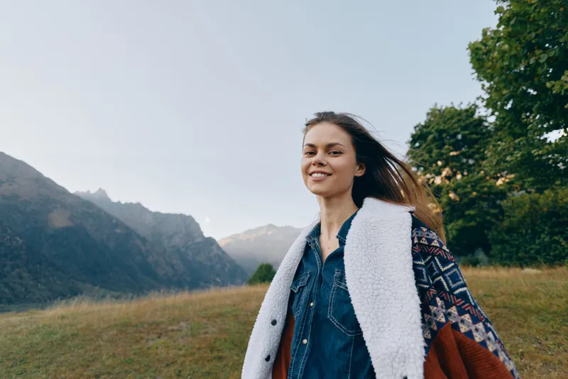 Woman smiling in mountains outdoors nature portrait, young traveler in warm jacket and patterned coat standing in grassy meadow with scenic landscape, sunlight and relaxed confident expression.