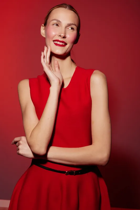 Smiling woman in red sleeveless dress poses with hand near face against red background. Fashion portrait with elegant makeup and confident expression in studio lighting.