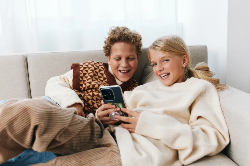 Two smiling children cuddle on a sofa, sharing a smartphone as soft daylight enters from a window, creating a cozy, candid moment of playful togetherness and curiosity.
