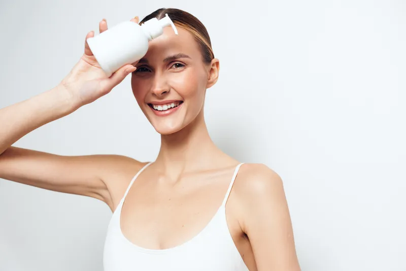A smiling woman applies lotion from a bottle to her hair in a bright studio, illustrating personal care routines, skincare and haircare, and a sense of confident wellness.