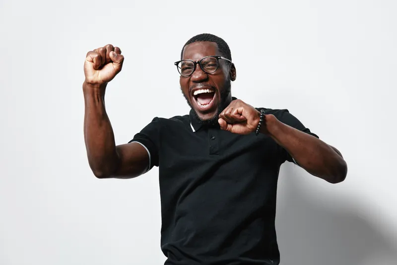 African American man with dark skin wearing glasses and black polo shirt celebrating with raised fists and joyful expression on white background. Excited, happy, success.